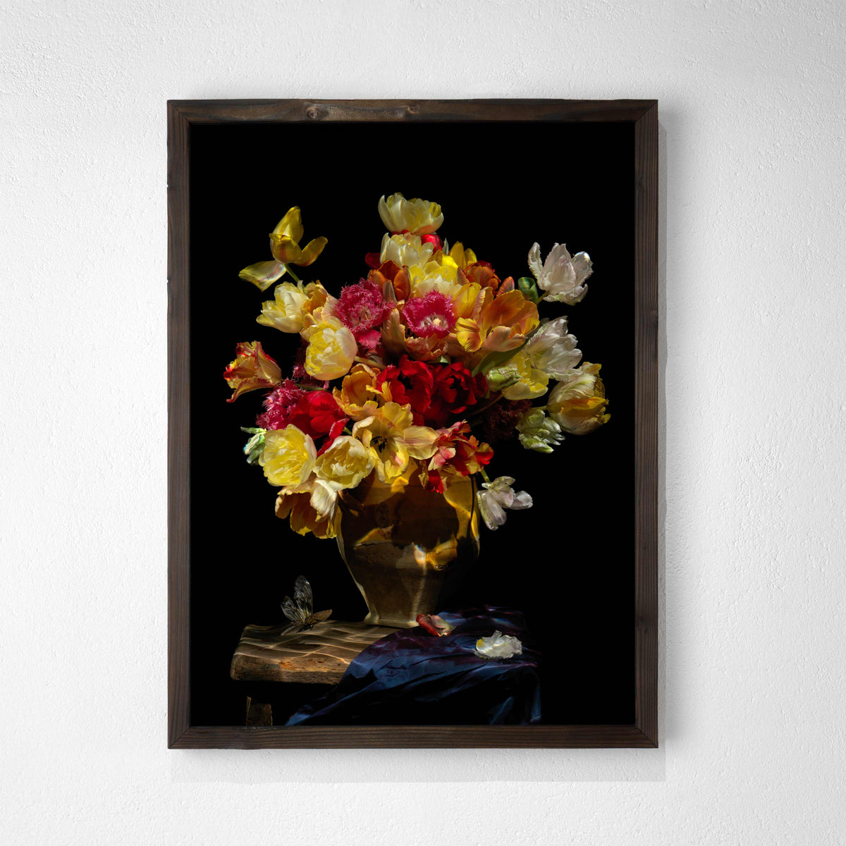 A framed still life art print featuring a vibrant bouquet of yellow, red, and white flowers in a gold vase against a black background, hung on a textured white wall.